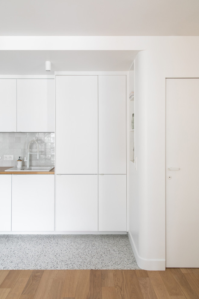 This is an example of a small contemporary single-wall eat-in kitchen in Paris with a single-bowl sink, beaded inset cabinets, white cabinets, wood benchtops, white splashback, ceramic splashback, white appliances, terrazzo floors, multi-coloured floor and beige benchtop.