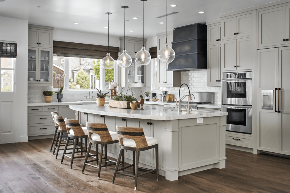 Inspiration for a large beach style l-shaped kitchen pantry in Orange County with an undermount sink, recessed-panel cabinets, beige cabinets, quartz benchtops, white splashback, porcelain splashback, stainless steel appliances, medium hardwood floors, with island, brown floor, white benchtop and vaulted.