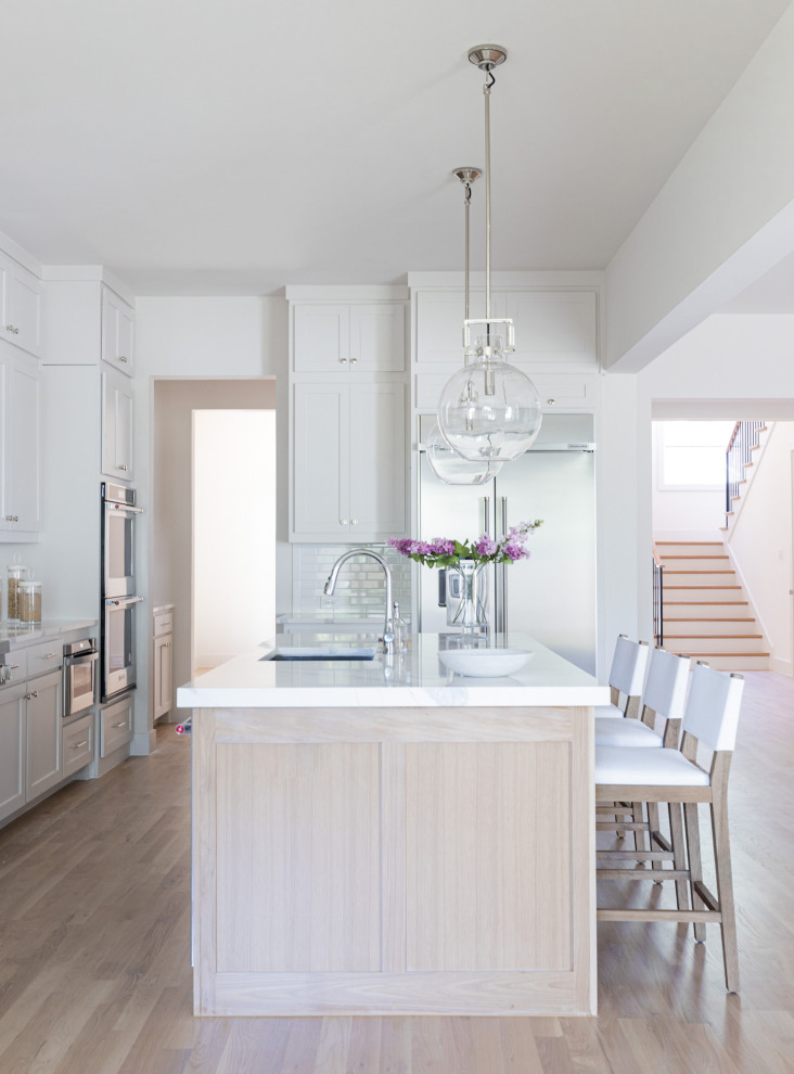 Expansive beach style single-wall kitchen/diner in Dallas with a submerged sink, shaker cabinets, white cabinets, engineered stone countertops, white splashback, porcelain splashback, stainless steel appliances, light hardwood flooring, an island, brown floors and white worktops.