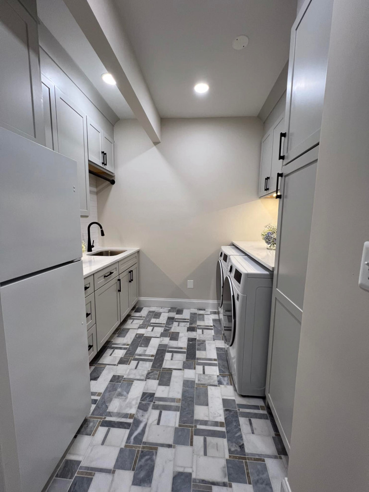 ワシントンD.C.にある高級な中くらいなコンテンポラリースタイルのおしゃれな洗濯室 (ll型、アンダーカウンターシンク、シェーカースタイル扉のキャビネット、グレーのキャビネット、クオーツストーンカウンター、白いキッチンパネル、セメントタイルのキッチンパネル、グレーの壁、大理石の床、左右配置の洗濯機・乾燥機、白い床、白いキッチンカウンター) の写真