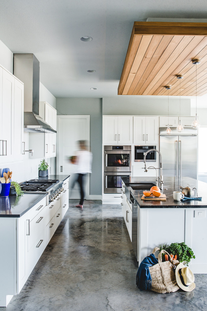 Kitchen - mid-sized transitional l-shaped concrete floor kitchen idea in Austin with a farmhouse sink, shaker cabinets, white cabinets, stainless steel appliances, an island, white backsplash, quartzite countertops, porcelain backsplash and black countertops