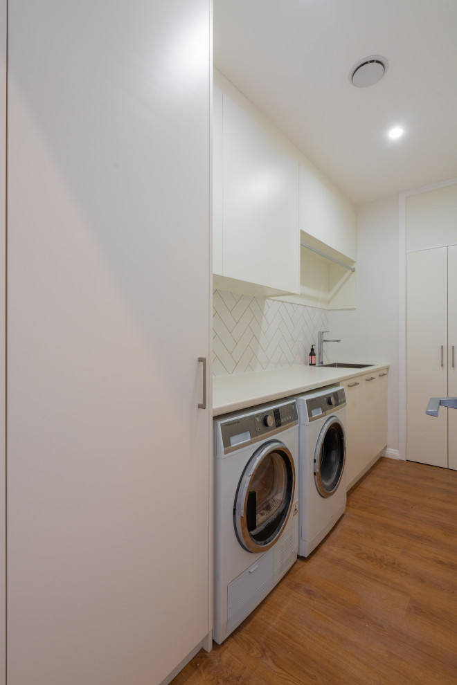 Design ideas for a mid-sized transitional single-wall dedicated laundry room in Auckland with a single-bowl sink, white cabinets, laminate benchtops, white splashback, subway tile splashback, white walls, laminate floors, a side-by-side washer and dryer, brown floor and white benchtop.