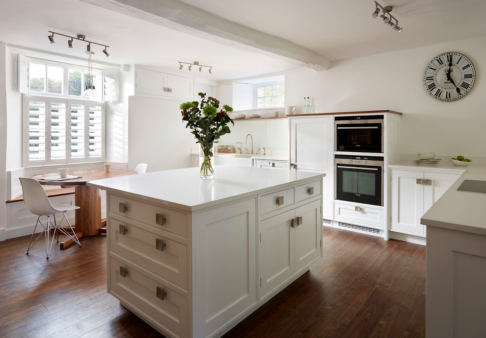 This is an example of a mid-sized contemporary eat-in kitchen in Buckinghamshire with shaker cabinets, white cabinets, quartzite benchtops, with island, an undermount sink, white splashback, glass sheet splashback, black appliances and dark hardwood floors.