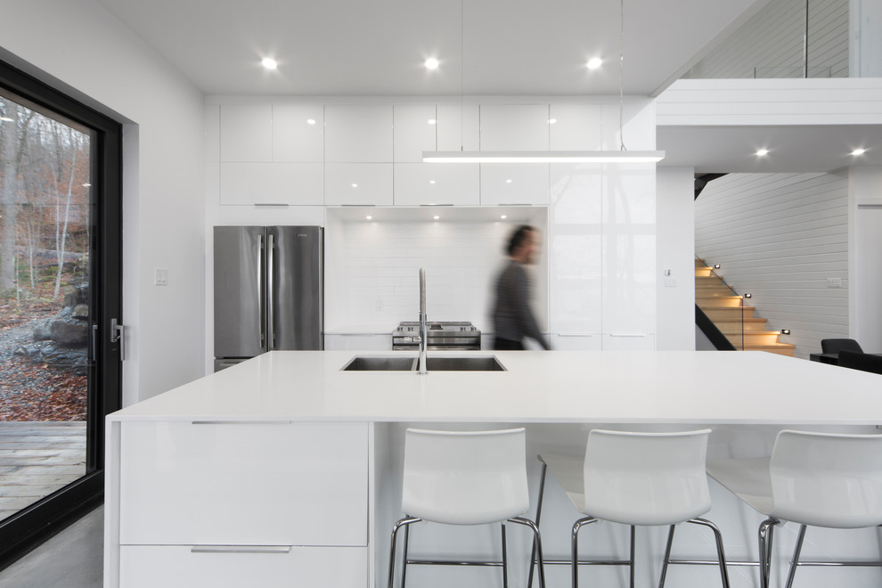 Inspiration for a mid-sized contemporary kitchen in Montreal with flat-panel cabinets, white cabinets, quartz benchtops, white splashback, ceramic splashback, stainless steel appliances, concrete floors, with island, a double-bowl sink and grey floor.