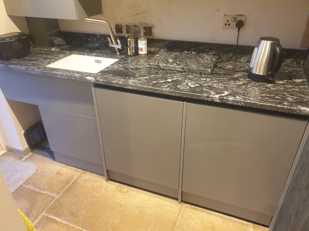 This is an example of a mid-sized contemporary l-shaped laundry room in West Midlands with flat-panel cabinets, granite benchtops, black splashback, engineered quartz splashback, black benchtop, a drop-in sink, black cabinets, travertine floors, beige floor and vaulted.