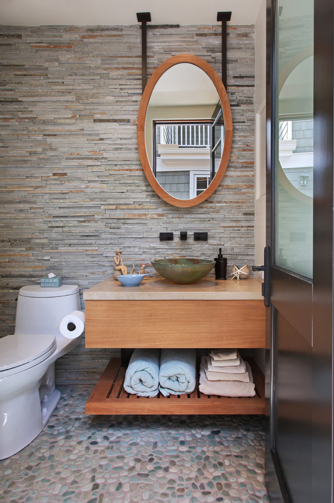 Photo of a mid-sized beach style bathroom in Orange County with a vessel sink, flat-panel cabinets, medium wood cabinets, a one-piece toilet, gray tile, stone tile, pebble tile floors and grey walls.