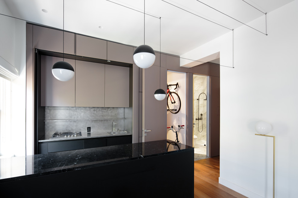 Photo of a small contemporary kitchen in New York with a drop-in sink, glass-front cabinets, grey cabinets, marble benchtops, metallic splashback, stainless steel appliances, medium hardwood floors, with island, beige floor, black benchtop and exposed beam.