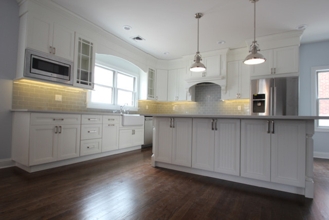 Example of a mid-sized transitional l-shaped brown floor and dark wood floor eat-in kitchen design with a farmhouse sink, recessed-panel cabinets, white cabinets, quartzite countertops, gray backsplash, ceramic backsplash, stainless steel appliances and an island