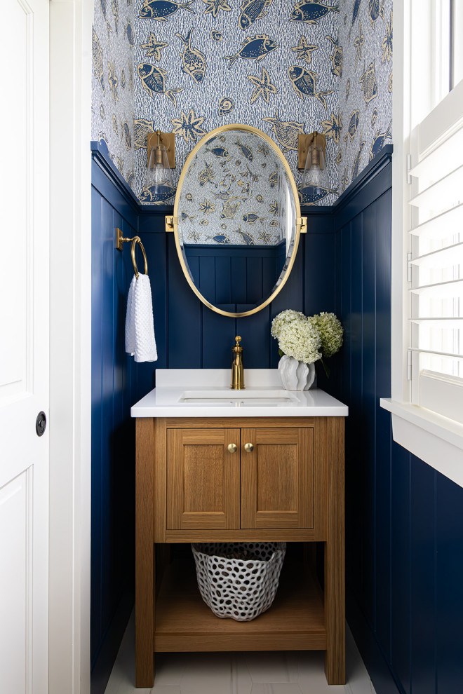 Small beach style wainscoting powder room photo in Boston with shaker cabinets, medium tone wood cabinets, blue walls, an undermount sink, quartz countertops, white countertops and a freestanding vanity