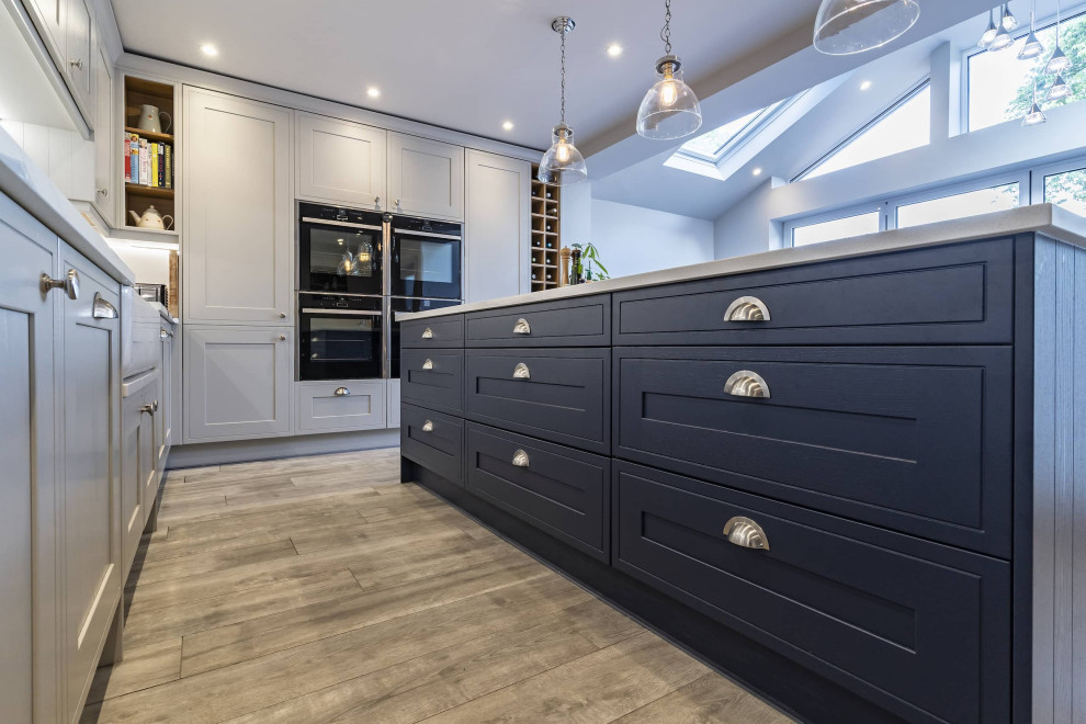 Photo of a medium sized traditional l-shaped open plan kitchen in Oxfordshire with a double-bowl sink, shaker cabinets, white cabinets, quartz worktops, white splashback, ceramic splashback, black appliances, ceramic flooring, an island, brown floors, white worktops and feature lighting.