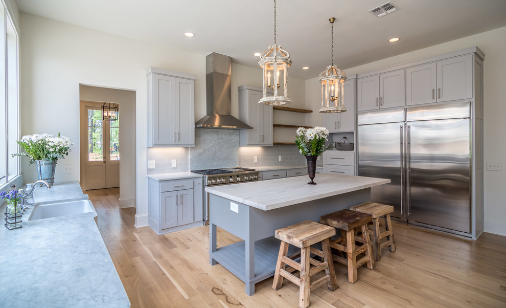 This is an example of a large country kitchen in New Orleans with a farmhouse sink, shaker cabinets, grey splashback, stainless steel appliances, light hardwood floors and with island.