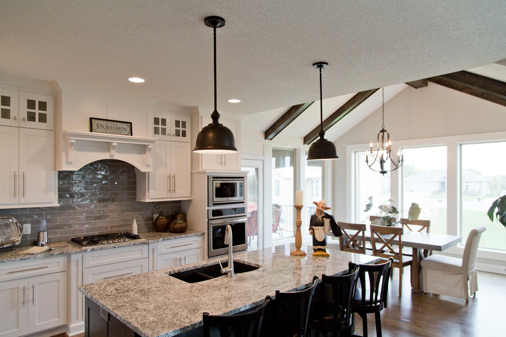 This is an example of a large country l-shaped open plan kitchen in Kansas City with an undermount sink, shaker cabinets, white cabinets, granite benchtops, grey splashback, ceramic splashback, stainless steel appliances, medium hardwood floors, with island, brown floor and beige benchtop.