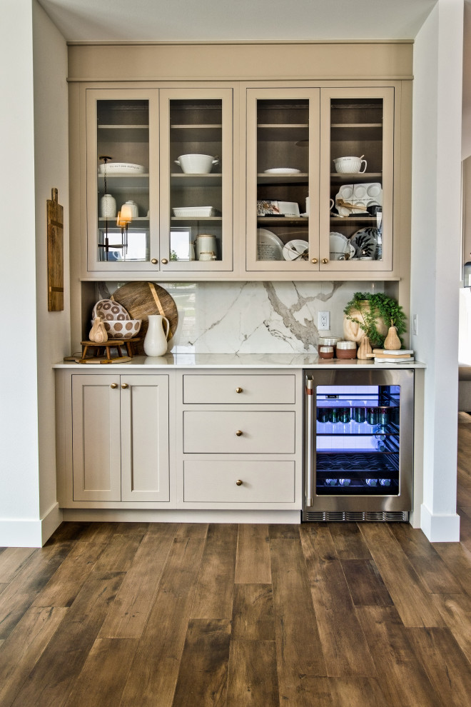Inspiration for a traditional home bar in Other with recessed-panel cabinets, beige cabinets, solid surface benchtops, white splashback, stone slab splashback, medium hardwood floors, brown floor and white benchtop.