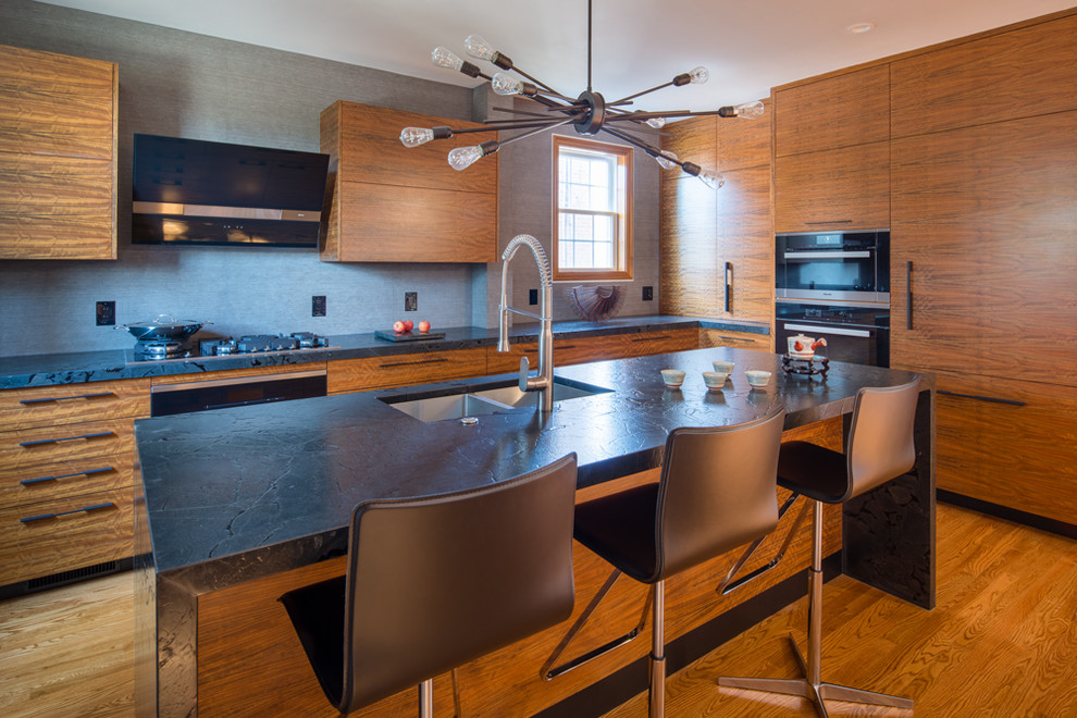 Photo of a mid-sized modern l-shaped eat-in kitchen in DC Metro with a double-bowl sink, flat-panel cabinets, medium wood cabinets, soapstone benchtops, beige splashback, cement tile splashback, stainless steel appliances, light hardwood floors, with island, beige floor and black benchtop.
