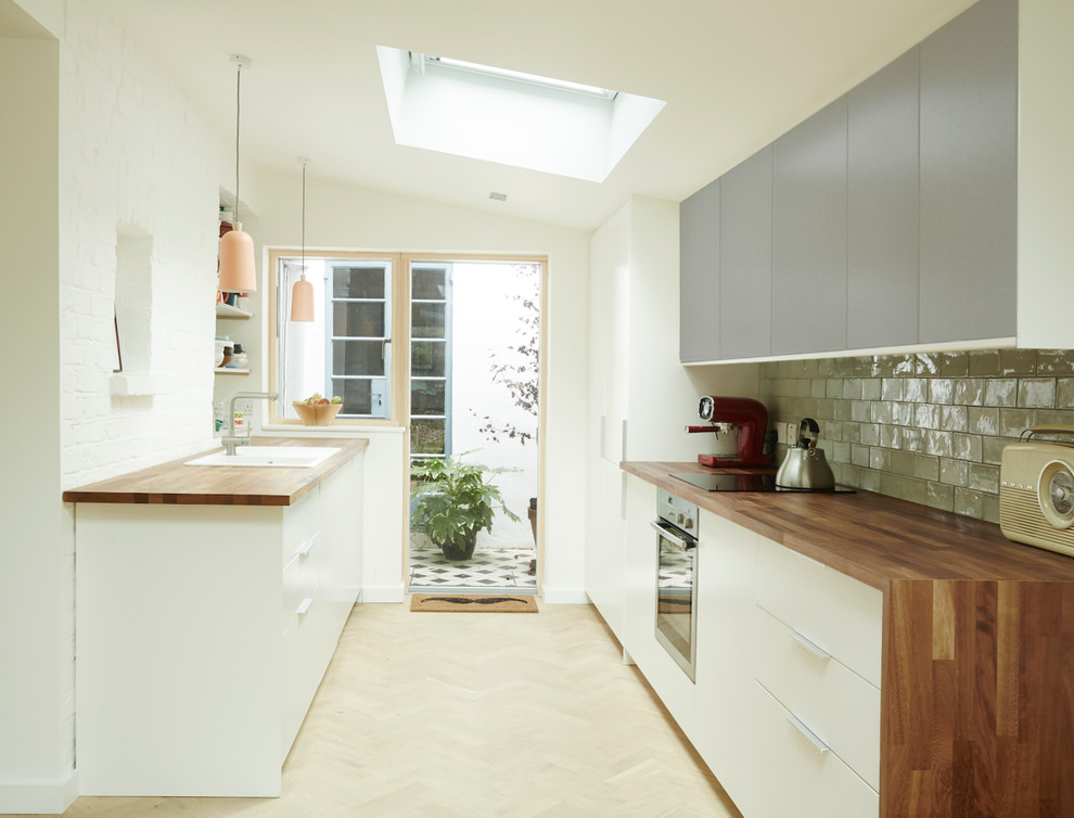 This is an example of a mid-sized contemporary galley kitchen in London with flat-panel cabinets, grey cabinets, wood benchtops, green splashback, ceramic splashback, stainless steel appliances, light hardwood floors and a drop-in sink.