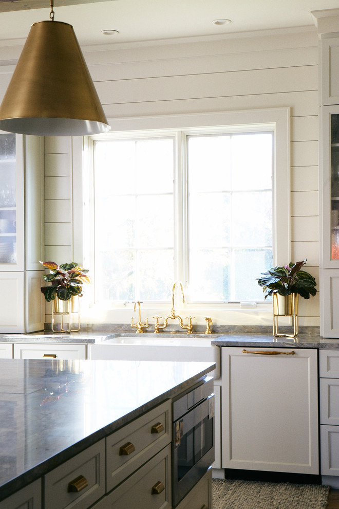 Large transitional l-shaped medium tone wood floor and brown floor eat-in kitchen photo with a farmhouse sink, raised-panel cabinets, white cabinets, marble countertops, gray backsplash, marble backsplash, stainless steel appliances, an island and gray countertops