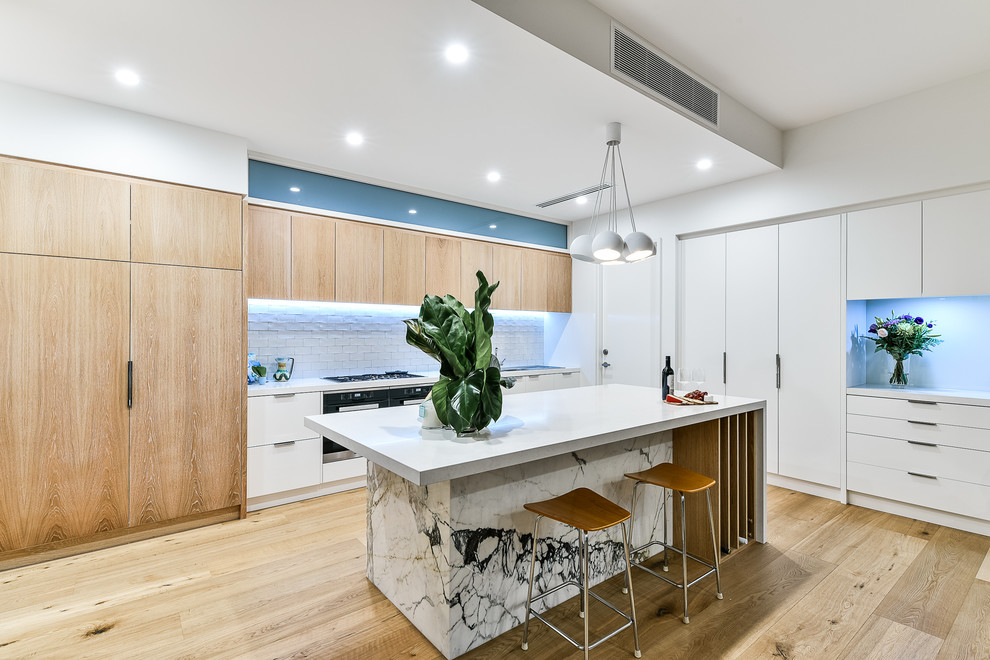 This is an example of a contemporary galley kitchen in Perth with flat-panel cabinets, white cabinets, white splashback, stainless steel appliances, light hardwood floors and with island.