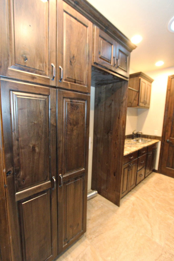 Inspiration for a large traditional galley dedicated laundry room in Boise with an undermount sink, raised-panel cabinets, dark wood cabinets, granite benchtops, beige walls, travertine floors and a stacked washer and dryer.