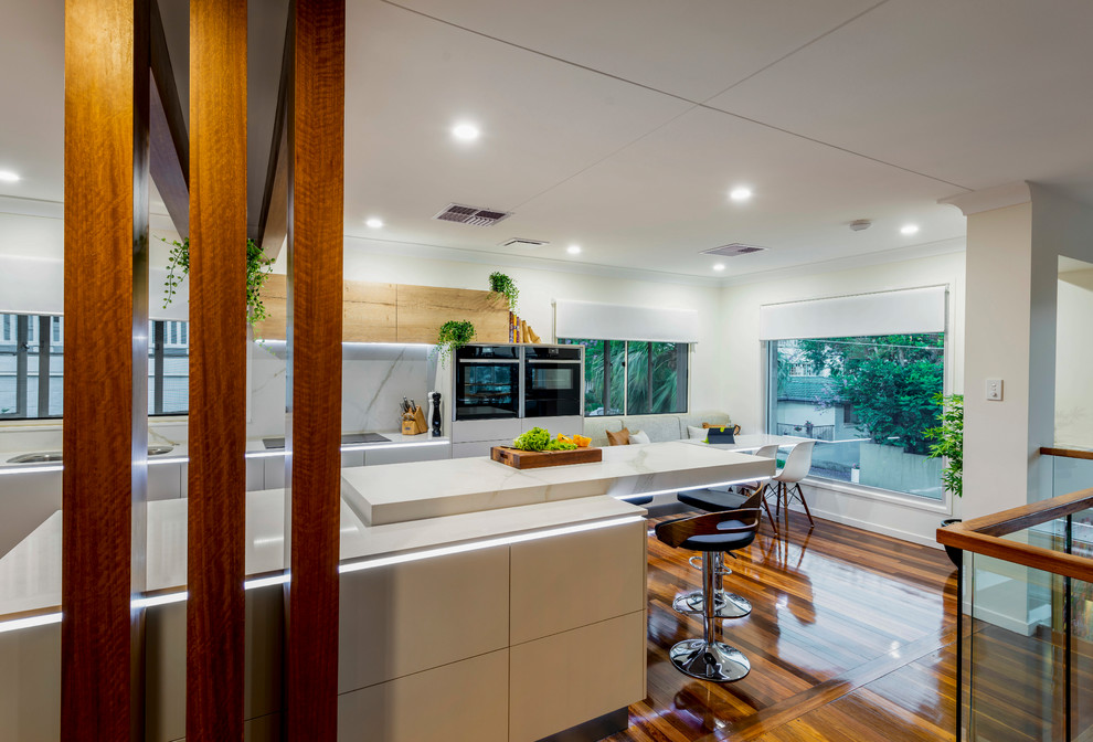 Photo of a large contemporary l-shaped eat-in kitchen in Brisbane with an undermount sink, flat-panel cabinets, beige cabinets, quartz benchtops, white splashback, stone slab splashback, black appliances, medium hardwood floors, with island, multi-coloured floor and yellow benchtop.