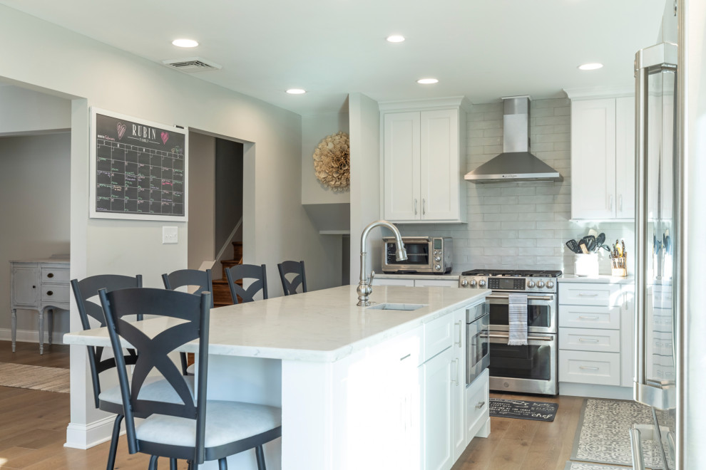 Mid-sized traditional l-shaped open plan kitchen in Philadelphia with an undermount sink, recessed-panel cabinets, white cabinets, quartzite benchtops, white splashback, porcelain splashback, stainless steel appliances, medium hardwood floors, with island, brown floor and beige benchtop.