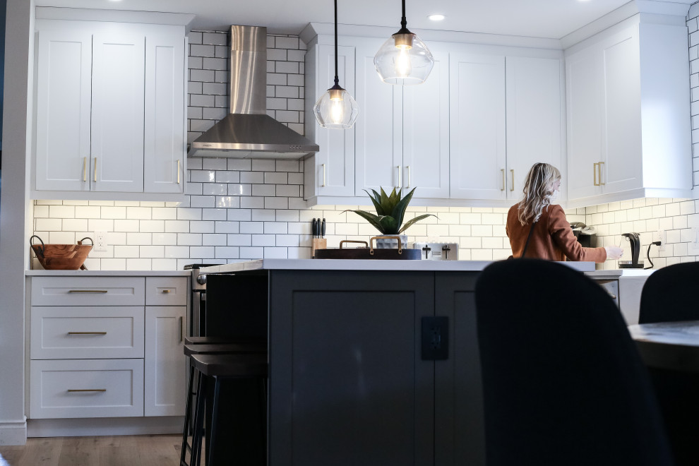 Small l-shaped light wood floor eat-in kitchen photo with a farmhouse sink, shaker cabinets, white cabinets, quartz countertops, white backsplash, porcelain backsplash, stainless steel appliances, an island and white countertops