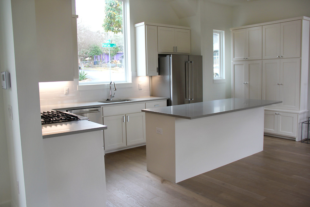 This is an example of a mid-sized modern u-shaped open plan kitchen in Austin with an undermount sink, shaker cabinets, white cabinets, quartz benchtops, white splashback, subway tile splashback, stainless steel appliances, medium hardwood floors, with island, brown floor and grey benchtop.