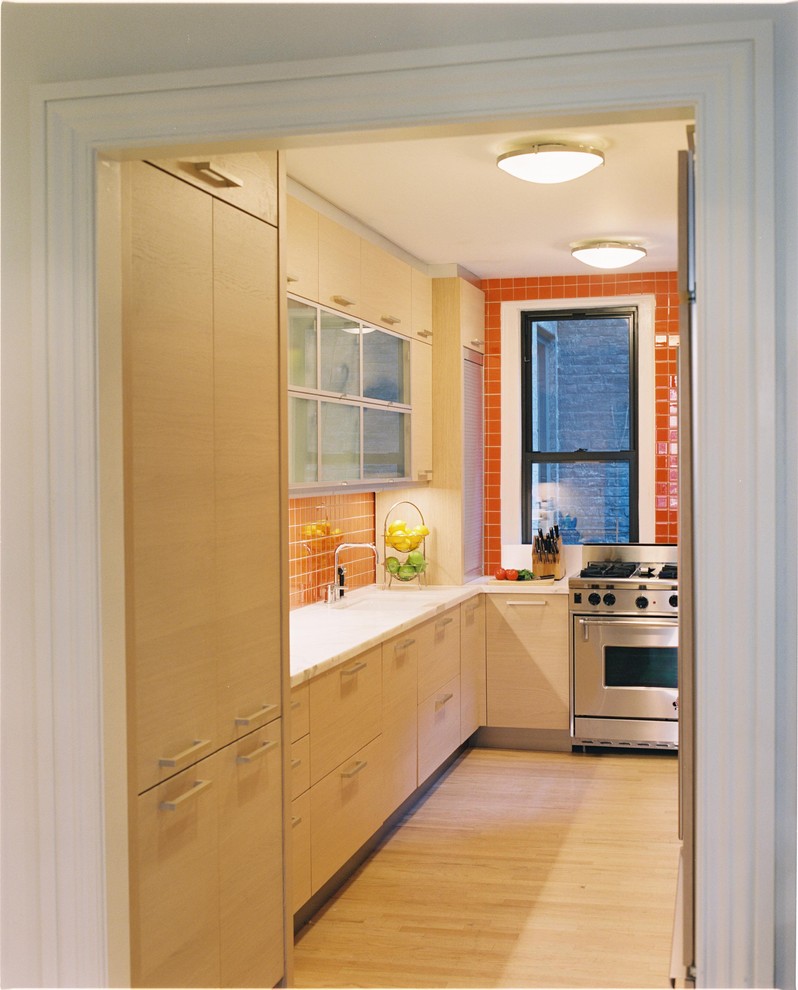 This is an example of a modern separate kitchen in New York with glass-front cabinets, stainless steel appliances, light wood cabinets and orange splashback.