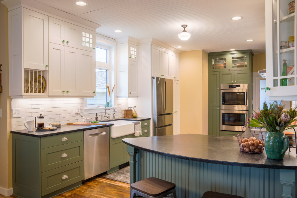 Photo of a country u-shaped eat-in kitchen in Minneapolis with a farmhouse sink, shaker cabinets, green cabinets, white splashback, subway tile splashback, stainless steel appliances, a peninsula, brown floor and black benchtop.
