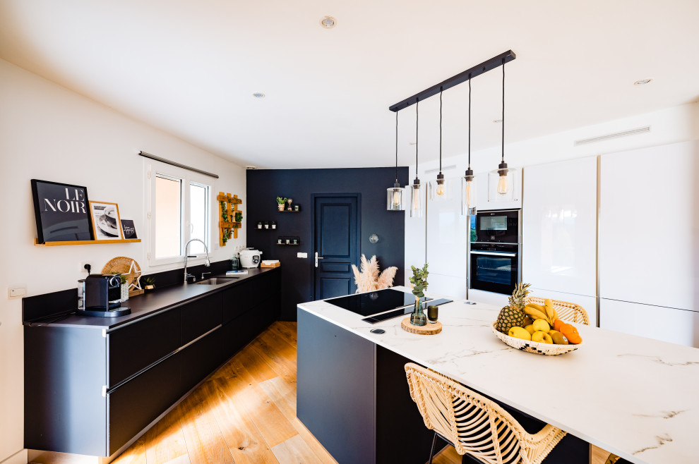 This is an example of a large contemporary u-shaped open plan kitchen in Other with a submerged sink, flat-panel cabinets, white cabinets, marble worktops, black splashback, marble splashback, black appliances, light hardwood flooring, an island, beige floors and white worktops.