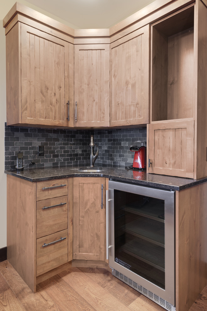This is an example of a small country l-shaped wet bar in Vancouver with an undermount sink, beaded inset cabinets, medium wood cabinets, granite benchtops, grey splashback, stone tile splashback, medium hardwood floors, brown floor and black benchtop.
