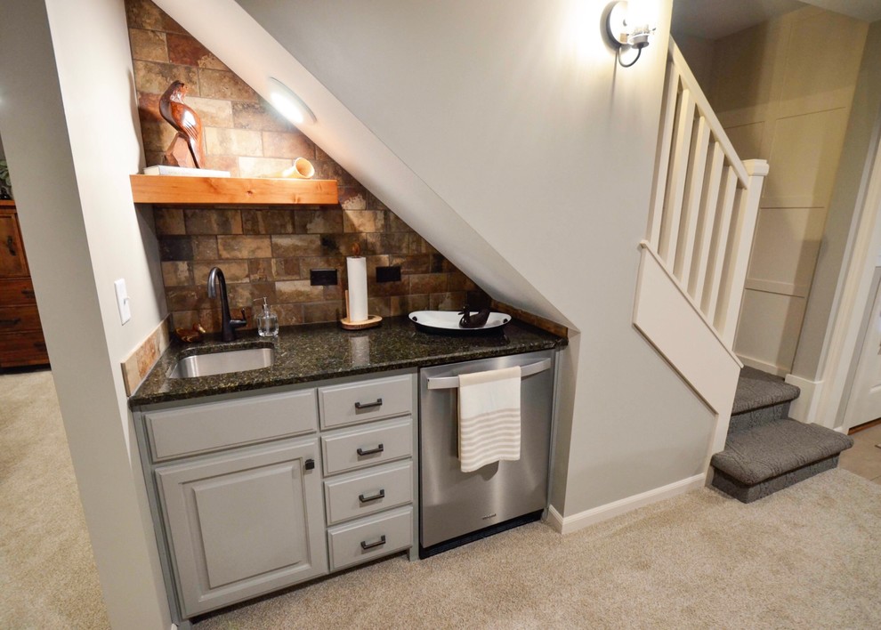 Photo of a small traditional single-wall wet bar in Kansas City with an undermount sink, raised-panel cabinets, grey cabinets, granite benchtops, brown splashback, stone tile splashback, carpet, beige floor and black benchtop.