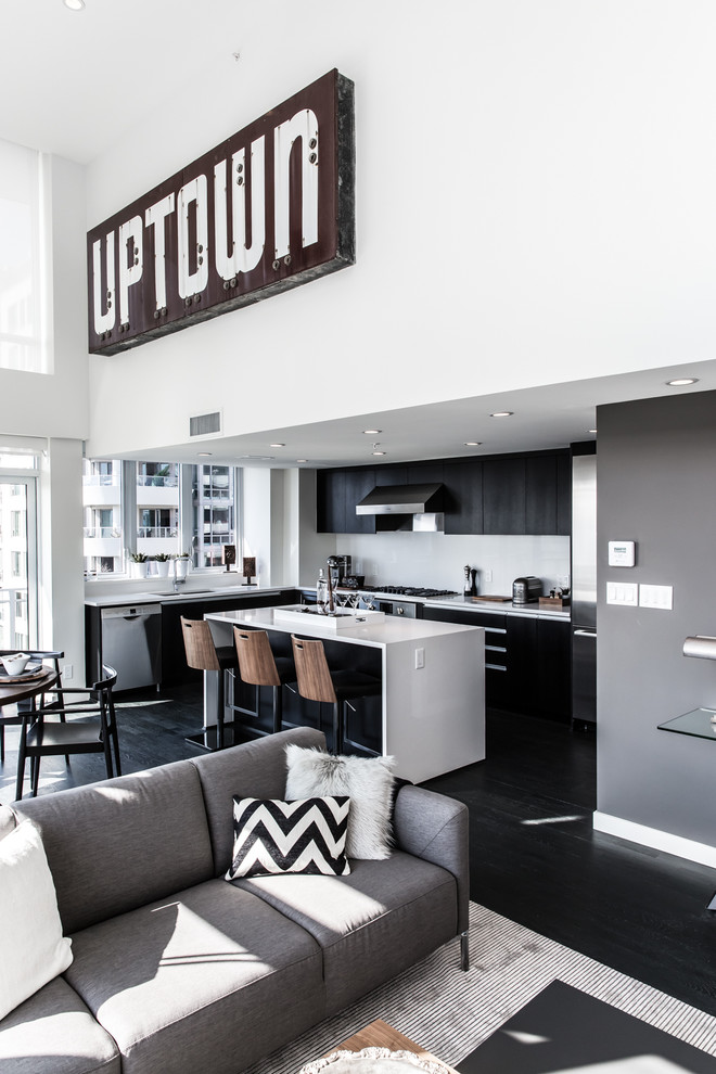 Mid-sized eclectic l-shaped open plan kitchen in Vancouver with flat-panel cabinets, black cabinets, solid surface benchtops, stainless steel appliances, dark hardwood floors, with island, black floor and grey splashback.