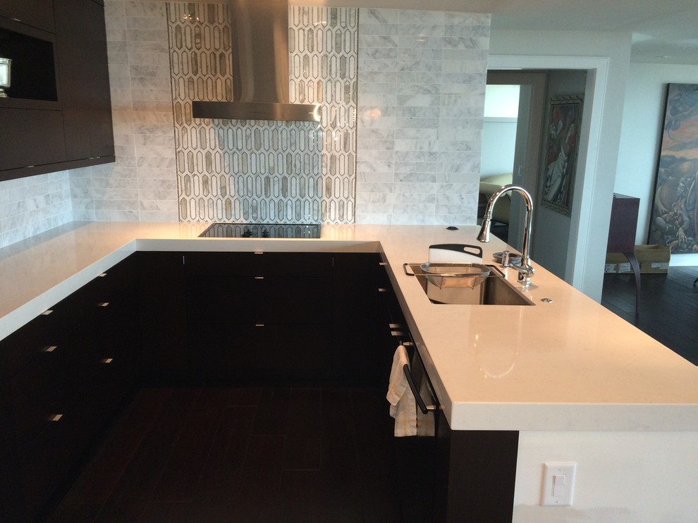 Mid-sized midcentury u-shaped open plan kitchen in Atlanta with flat-panel cabinets, dark wood cabinets, an undermount sink, solid surface benchtops, white splashback, marble splashback, black appliances, brown floor and with island.