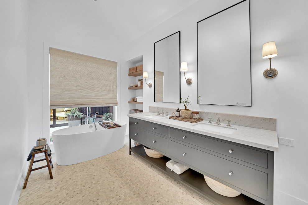 Photo of a beach style bathroom in Orange County with grey cabinets, a freestanding tub, gray tile, pebble tile floors, an undermount sink, grey benchtops, a built-in vanity, marble benchtops, multi-coloured floor and a double vanity.