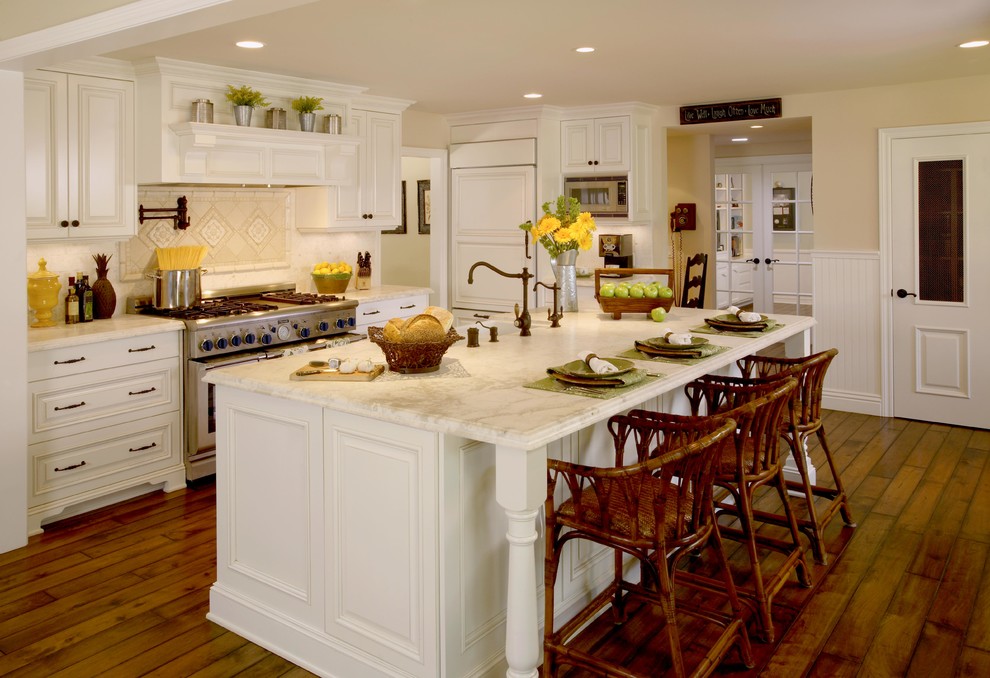 This is an example of a large transitional l-shaped kitchen in Orange County with a farmhouse sink, raised-panel cabinets, white cabinets, white splashback, marble benchtops, ceramic splashback, panelled appliances, dark hardwood floors, with island and brown floor.