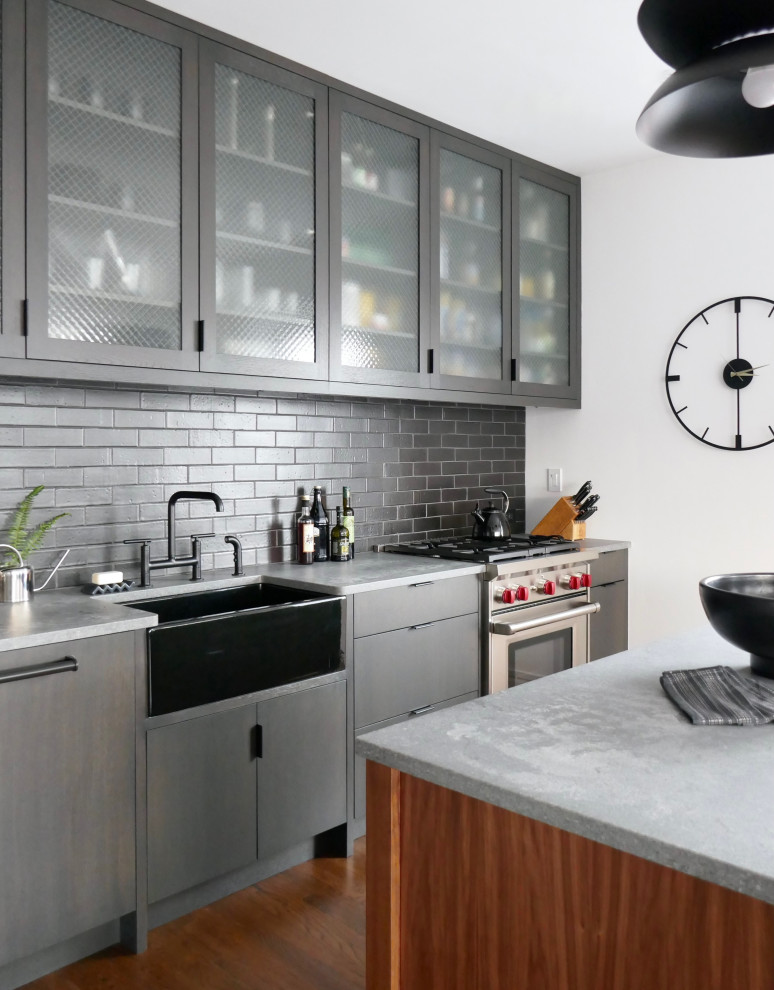 This is an example of an expansive modern l-shaped eat-in kitchen in New York with a farmhouse sink, glass-front cabinets, grey cabinets, quartz benchtops, black splashback, brick splashback, stainless steel appliances, medium hardwood floors, with island, brown floor and grey benchtop.