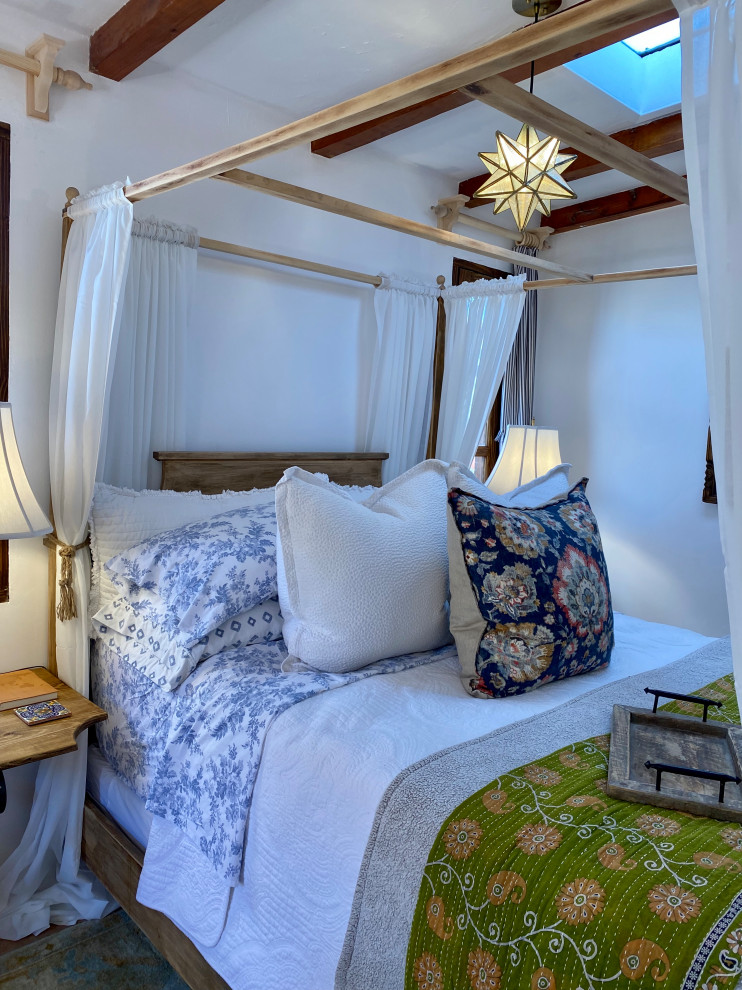 На фото: гостевая спальня среднего размера, (комната для гостей), в светлых тонах в средиземноморском стиле с белыми стенами, полом из керамогранита, угловым камином, фасадом камина из штукатурки, оранжевым полом и балками на потолке с