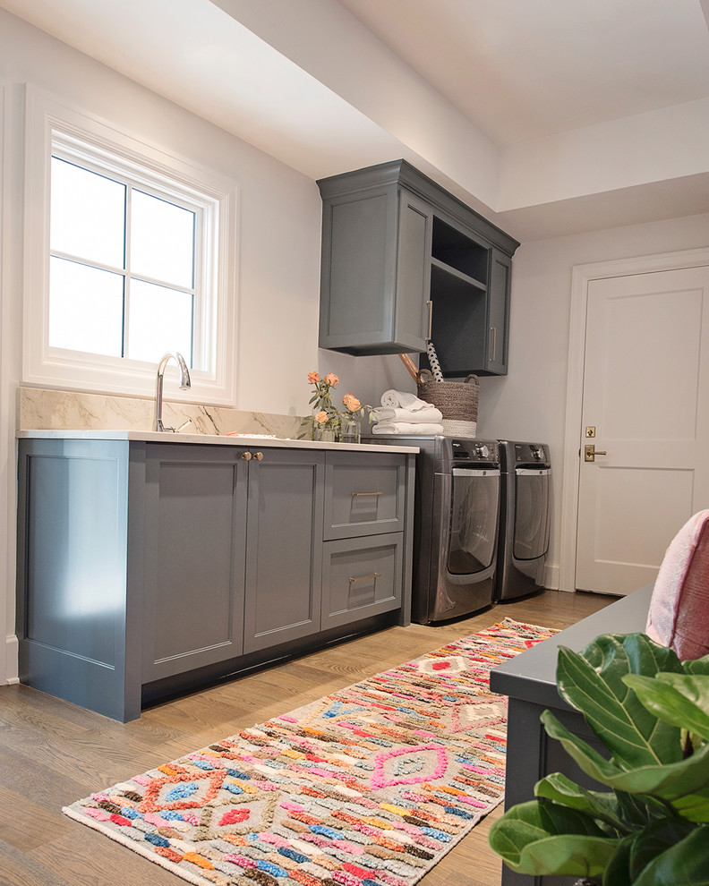 Photo of a large eclectic l-shaped utility room in Austin with recessed-panel cabinets, grey cabinets, white walls, light hardwood floors, a side-by-side washer and dryer and brown floor.