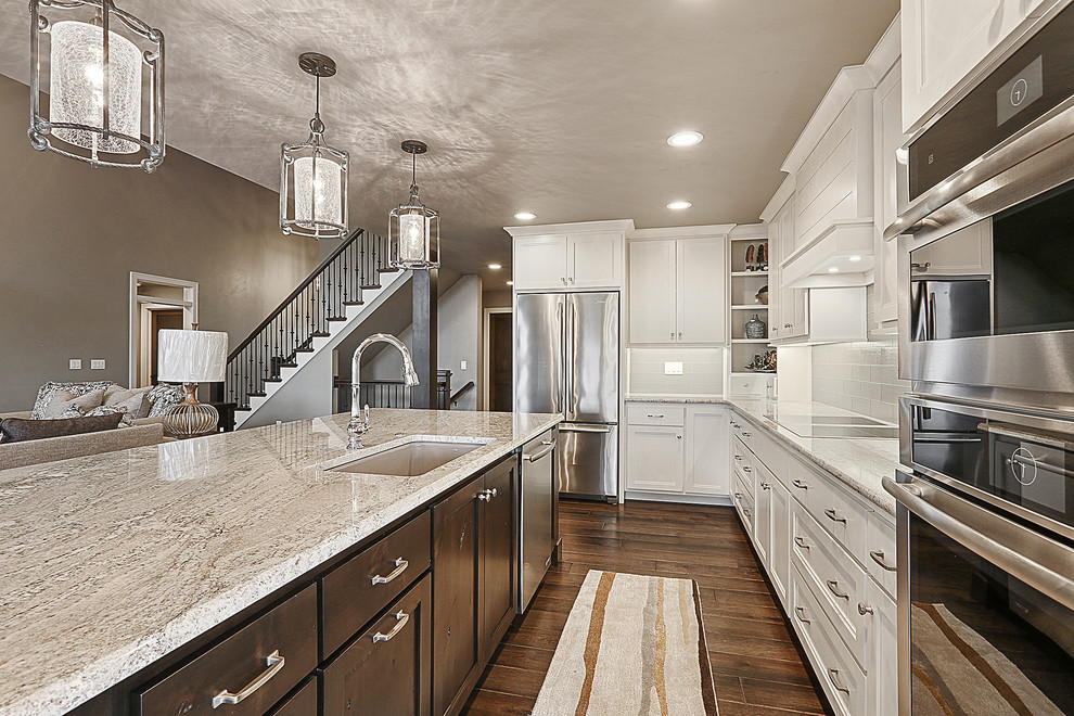 Example of a dark wood floor and brown floor kitchen design with an undermount sink, shaker cabinets, granite countertops, gray backsplash, glass tile backsplash, stainless steel appliances and an island