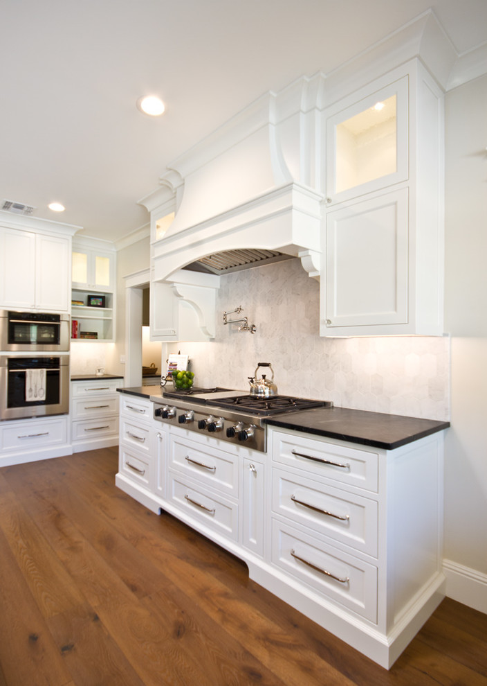 Large transitional u-shaped medium tone wood floor eat-in kitchen photo in Phoenix with a farmhouse sink, recessed-panel cabinets, white cabinets, marble countertops, white backsplash, stone tile backsplash, stainless steel appliances and an island