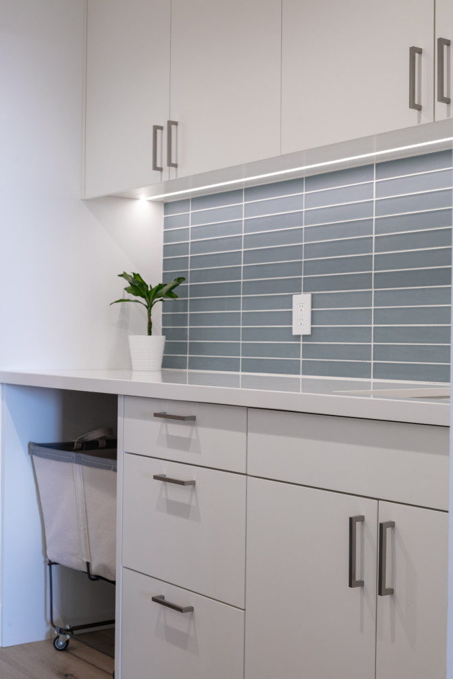 Design ideas for a mid-sized modern galley dedicated laundry room in Orange County with an undermount sink, flat-panel cabinets, white cabinets, glass benchtops, blue splashback, glass tile splashback, blue walls, medium hardwood floors, beige floor and white benchtop.