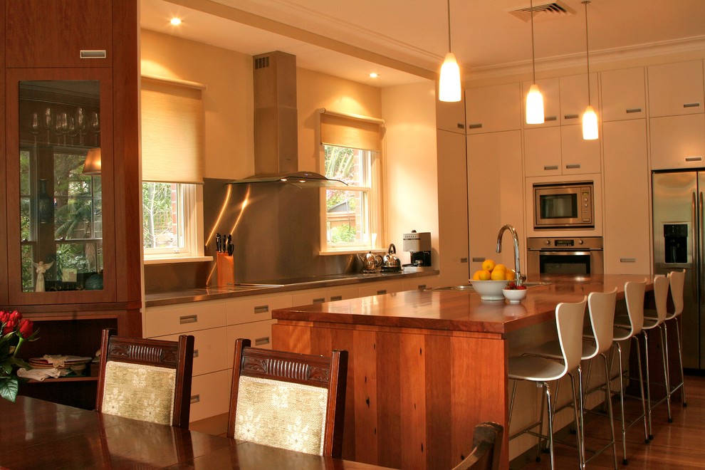 Large transitional l-shaped eat-in kitchen in Sydney with a drop-in sink, flat-panel cabinets, white cabinets, wood benchtops, metallic splashback, stainless steel appliances, medium hardwood floors and with island.