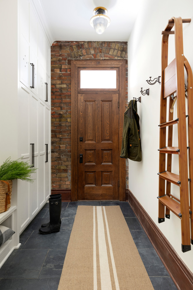 На фото: тамбур с белыми стенами, одностворчатой входной дверью, входной дверью из дерева среднего тона, серым полом и кирпичными стенами