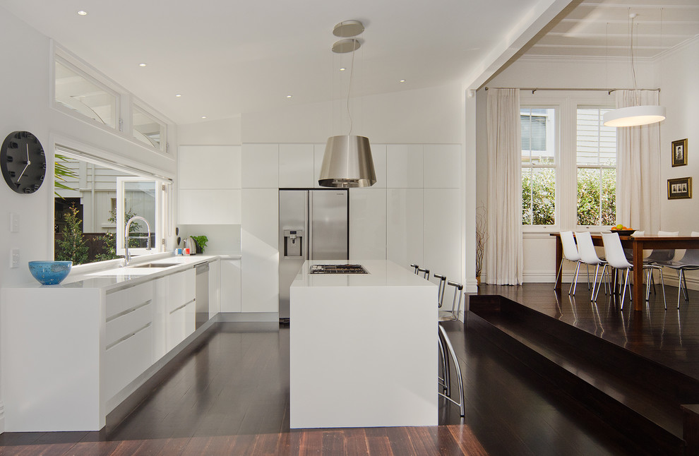 Photo of a mid-sized contemporary l-shaped eat-in kitchen in Auckland with an undermount sink, flat-panel cabinets, white cabinets, solid surface benchtops, stainless steel appliances, dark hardwood floors, with island and white splashback.