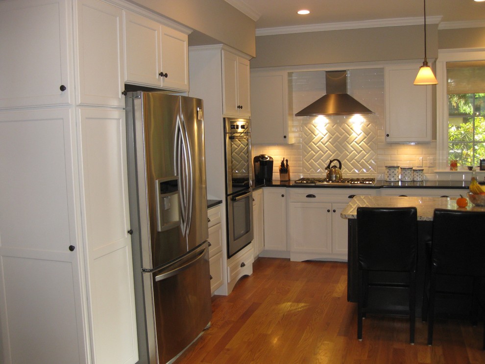 Transitional l-shaped medium tone wood floor eat-in kitchen photo in Boston with an undermount sink, shaker cabinets, white cabinets, granite countertops, white backsplash, ceramic backsplash, stainless steel appliances, an island and black countertops