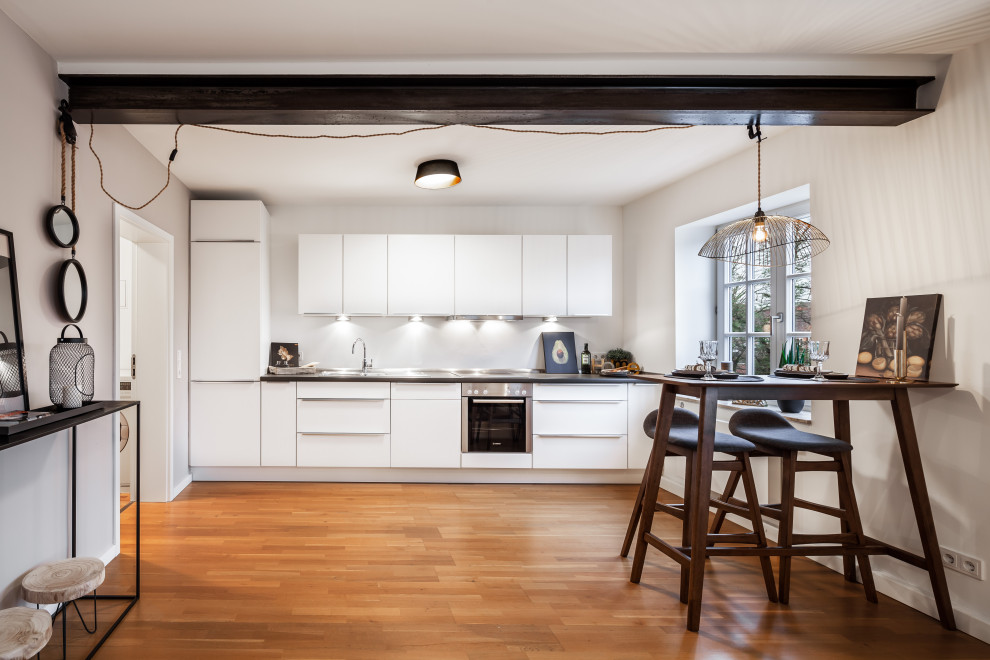 Einzeilige Moderne Wohnküche mit flächenbündigen Schrankfronten, weißen Schränken, Küchengeräten aus Edelstahl, braunem Holzboden, braunem Boden, grauer Arbeitsplatte und freigelegten Dachbalken in Hamburg