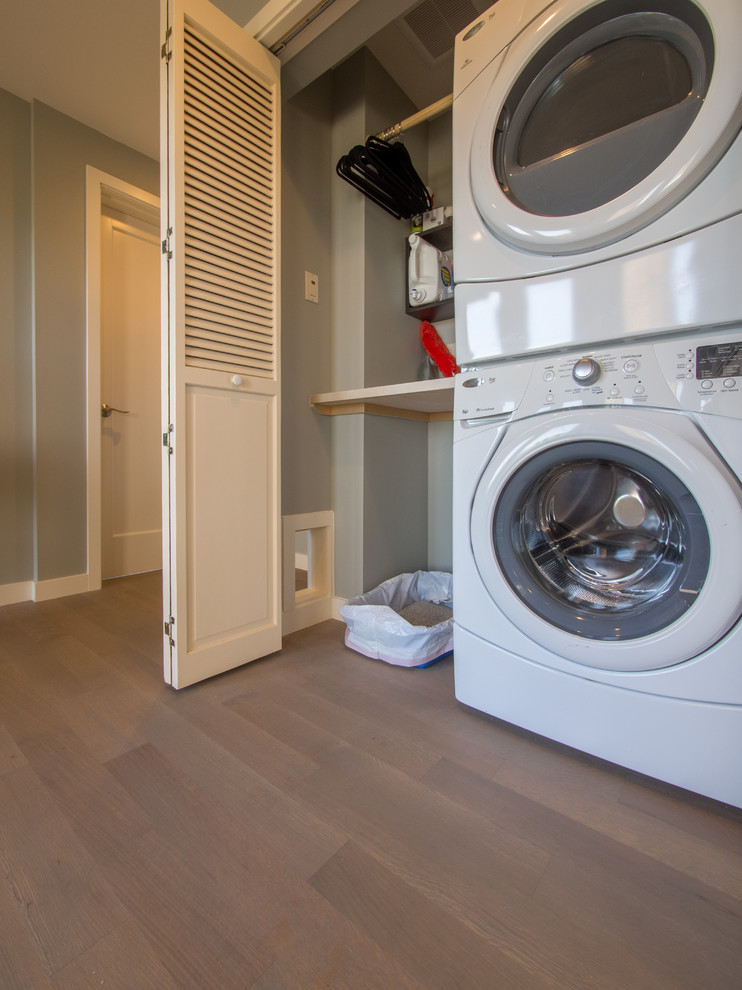 This is an example of a small transitional single-wall laundry cupboard in Seattle with solid surface benchtops, medium hardwood floors, a stacked washer and dryer and grey walls.