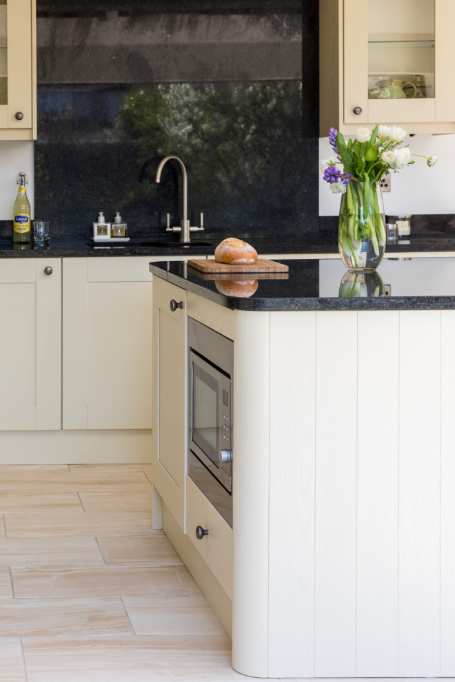 This is an example of a large classic cream and black l-shaped open plan kitchen in West Midlands with a double-bowl sink, glass-front cabinets, white cabinets, granite worktops, black splashback, laminate floors, an island, black worktops and exposed beams.