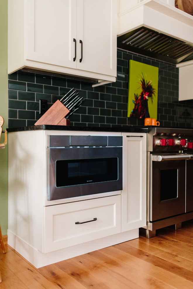 Large transitional galley light wood floor and brown floor open concept kitchen photo with a farmhouse sink, recessed-panel cabinets, white cabinets, granite countertops, green backsplash, glass tile backsplash, paneled appliances and an island