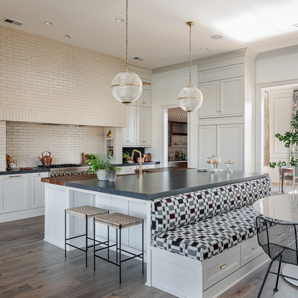 Transitional l-shaped eat-in kitchen in Los Angeles with a farmhouse sink, recessed-panel cabinets, white cabinets, beige splashback, brick splashback, stainless steel appliances, dark hardwood floors, with island, brown floor and grey benchtop.